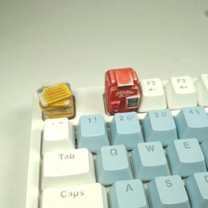 May include: A white mechanical keyboard with light blue and white keycaps. Custom keycaps shaped like a stack of toast and a red retro-style radio are on the keyboard. The keyboard has a variety of function keys and letter keys.