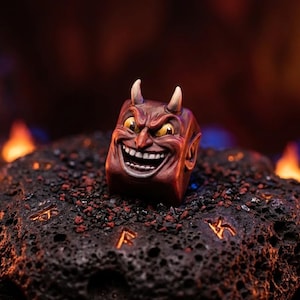 May include: A square-shaped artisan keycap featuring a sculpted, grinning devil face with pointed ears, two small horns, and yellow eyes. The keycap is painted in shades of deep red and brown, showcasing a detailed, expressive facial expression.