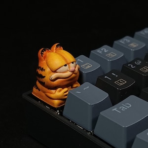 May include: A custom orange Garfield keycap on a mechanical keyboard. The keycap features a detailed, hand-painted design of the cartoon cat. The keyboard has gray and black keys.