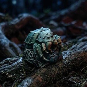May include: A sculpted artisan keycap featuring a detailed spider with multiple black eyes, a textured green and grey body, and intricate web patterns along the sides. The piece has a weathered finish with visible highlights and shadows.