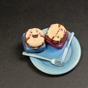 May include: Two whimsical, cartoon-style miniature cakes with frosting and decorative elements sit on a blue plate with a fork and knife. One cake has a smiling face, the other has floral accents. The cakes rest on small, colorful platforms.
