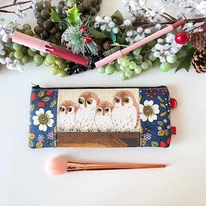 May include: A rectangular zippered pouch featuring a central panel with four illustrated owls perched on a branch, flanked by dark blue fabric with a floral and berry pattern. The pouch is accompanied by a pink lipstick, a pink pencil, and a makeup brush.
