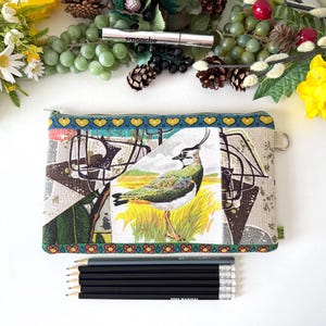 May include: A rectangular zipped pouch featuring a central illustration of a lapwing bird in a grassy field, surrounded by a geometric pattern in shades of green, brown, and cream. The pouch has a teal border with yellow hearts and orange flowers.
