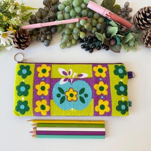 May include: A zippered pencil case with a floral design in green, yellow, and purple. The case features an apple-shaped design in the center. Pencils in various colors are below the case. Makeup and decorative elements are in the background.