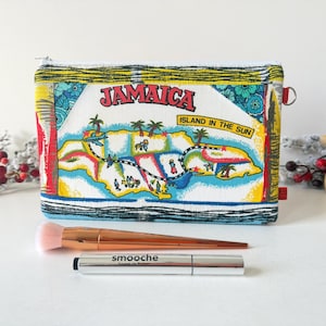 May include: A rectangular fabric zip pouch featuring a colourful map of Jamaica with the text JAMAICA ISLAND IN THE SUN. Below the pouch lies a rose gold makeup brush and a silver tube of Smooche lip gloss on a white surface.