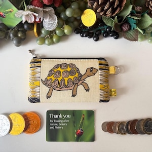 May include: A small, rectangular coin purse with a tortoise design. The purse is beige with a yellow and black patterned border. A small card with the text "Thank you" and various coins are also visible.