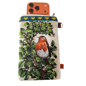 May include: A beige phone crossbody bag featuring a detailed illustration of a robin perched on a tree branch with green leaves. The top of the bag has a blue border with yellow hearts. A brown leather loop is attached to the side.