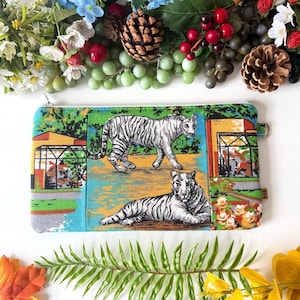 May include: A rectangular zipped pouch with a vibrant print of two white tigers in a zoo. The pouch has a teal, green, and orange colour scheme, with a small loop on the side. The background includes images of trees and buildings.