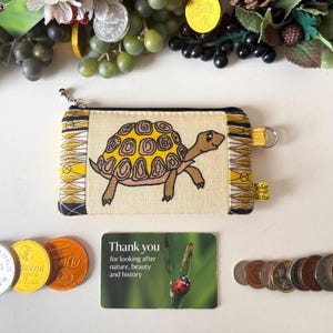 May include: A rectangular beige fabric coin purse featuring an embroidered yellow and brown turtle on the front. The purse has a black zipper, a silver metal ring on the side, and patterned black and yellow fabric panels on the left and right edges.