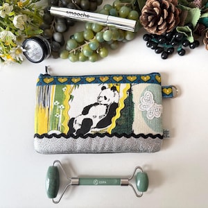 May include: A panda-themed zippered pouch with a silver base, a jade face roller, and makeup items. The pouch features a panda illustration, a butterfly applique, and a blue and yellow patterned border. The roller has a silver handle and green jade stones.