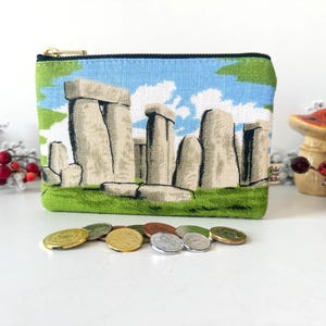 May include: A rectangular fabric zipper pouch featuring a woven graphic of Stonehenge with large grey stone monoliths set against a bright blue sky and green grass. The pouch has a gold-toned metal zipper closure along the top edge.