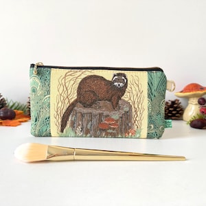 May include: A rectangular fabric zip pouch featuring a central illustration of a brown ferret perched on a tree stump, surrounded by green patterned side panels. The pouch has a black zip closure and a small gold-toned metal pull tab.