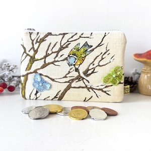 Op de afbeelding: Een rechthoekige beige stoffen portemonnee met een print van bruine boomtakken en een kleine gele en blauwe vogel. De portemonnee is versierd met een lichtblauwe vlinder van lovertjes en een groene vlinder van kralen, met een zilveren rits.