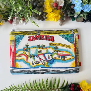 May include: Rectangular travel pouch featuring a colorful map of Jamaica with the text "JAMAICA ISLAND IN THE SUN." The pouch has a zipper closure and is trimmed with black and white stripes. Red tabs are on the sides.