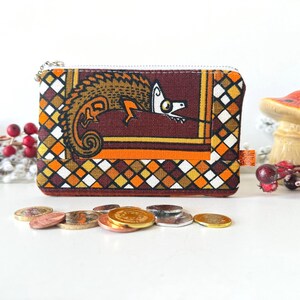 May include: A rectangular fabric coin purse featuring a graphic illustration of a stylised chameleon in shades of brown, orange, and yellow. The border has a diamond pattern in black, white, and orange. A small orange tag reads Under Over.
