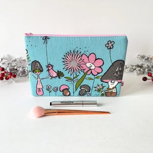 May include: A light blue zipped pouch featuring a whimsical illustration of pink flowers, small mushrooms, and a bird. The pouch has a pink zip and is displayed with a makeup brush and a silver tube labelled smooche.