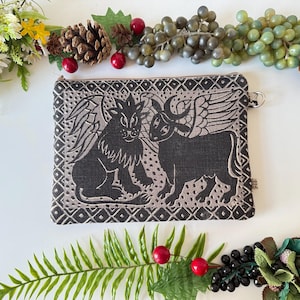 May include: Black and gray zippered pouch featuring winged lion and bull illustrations. The rectangular pouch has a decorative border and a small silver-colored ring attached to the side. The background is white with floral and berry accents.