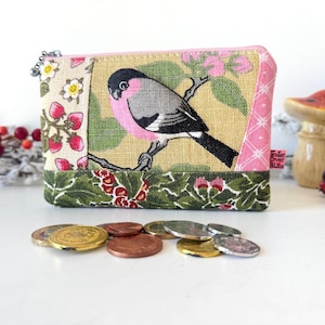 May include: A rectangular fabric coin purse featuring a central panel with a bullfinch bird on a branch, flanked by strawberry and pink patterned fabric sections. The bottom panel is green with a floral print and a small pink tag on the side.