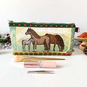 May include: A rectangular fabric zip pouch featuring a printed scene of a mare, a foal, and a grazing horse in a field. The pouch has a green border with a red and yellow floral pattern, a gold zip, and a small brown side tab.