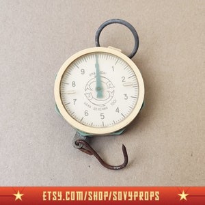 May include: A vintage cream-coloured hanging scale with a circular dial and a rusty hook. The dial features black numerals and a green graphic. The scale has a metal loop for hanging and a green frame. The scale measures up to 10 kg.