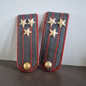 May include: Two black and red military shoulder boards with gold stars. The shoulder boards have three gold stars on each.