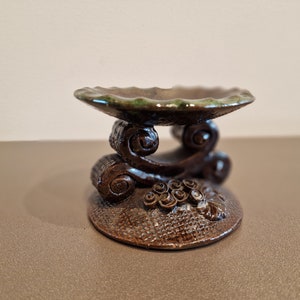 Pode incluir: Um pequeno pedestal de cerâmica marrom com uma parte superior esmaltada de verde. O pedestal tem um design decorativo de volutas e flores.