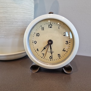 Può includere: Sveglia vintage bianca con numeri e lancette neri. L'orologio ha un quadrante in vetro ed è su un supporto bianco.