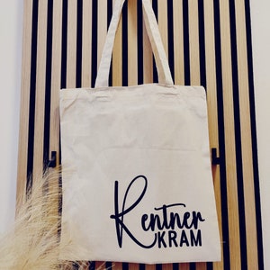 Könnte beinhalten: Eine weiße Stofftasche mit schwarzen Henkeln und dem Text "Rentner Kram" in schwarzer Kursivschrift.