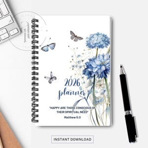 May include: A spiral-bound 2026 planner with a white cover featuring blue watercolor flowers and butterflies. The text "2026 planner" is in a script font, with a quote from Matthew 5:3. A black pen rests beside the planner.