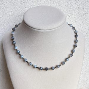 May include: A beaded necklace with gray and blue-gray stones and small black beads. The necklace is displayed on a white mannequin bust. The stones have a marbled appearance, and the necklace is strung on a dark cord.