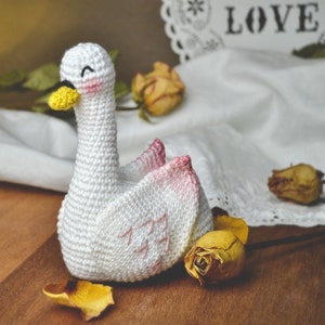 Op de afbeelding: Een witte gehaakte zwaan met roze accenten en zwarte ogen. De zwaan zit op een houten oppervlak met gedroogde bloemen.