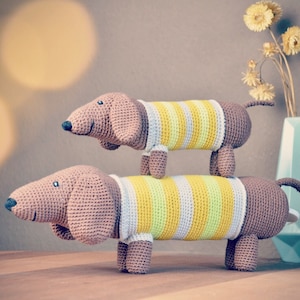 Könnte beinhalten: Zwei gehäkelte Dackel-Spielzeugtiere, eines auf dem anderen gestapelt. Beide Hunde tragen gestreifte Shirts mit gelben und weißen Streifen. Der obere Hund ist braun und der untere Hund ist hellbraun.
