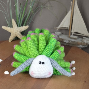 May include: A crocheted sea creature toy with a green and pink shell and a white face with black eyes. The toy is sitting on a wooden surface.