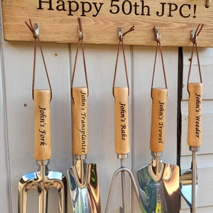 May include: A wooden sign with the text "Happy 50th JPC!" hangs above a set of five gardening tools. Each tool has a wooden handle and a metal head, labeled "John's Fork," "John's Transplanter," "John's Rake," "John's Trowel," and "John's Weeder."