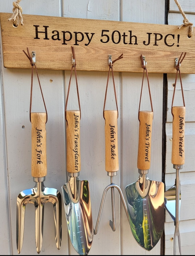 May include: A wooden sign with the text "Happy 50th JPC!" hangs above a set of five gardening tools. Each tool has a wooden handle and a metal head, labeled "John's Fork," "John's Transplanter," "John's Rake," "John's Trowel," and "John's Weeder."