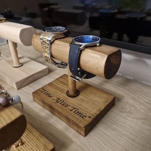 May include: Wooden watch stand with two watches, one with a gold band and one with a black band. The stand has the words "Time After Time" engraved on the base.