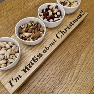 May include: A wooden serving board with the text "I'm nuts about Christmas!!" features four small white bowls filled with various nuts and dried cranberries. The board is made of light-colored wood and is set on a wooden surface.