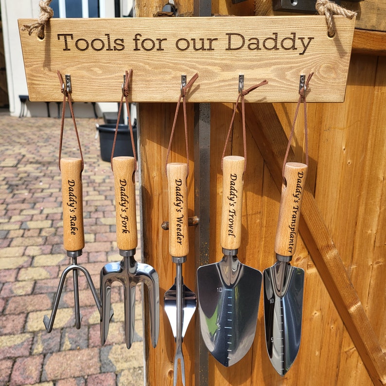 May include: A wooden sign with the text "Tools for our Daddy" hangs above a set of gardening tools. The tools include a rake, fork, weeder, trowel, and transplanter, each with a wooden handle and the words "Daddy's" followed by the tool name.