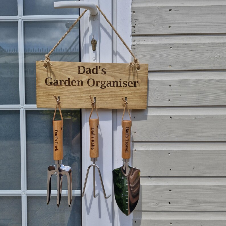 May include: Wooden sign that reads "Dad's Garden Organiser" with three garden tools hanging below. The tools include a fork, rake, and trowel, each with a wooden handle and leather strap. The sign is hanging on a door.