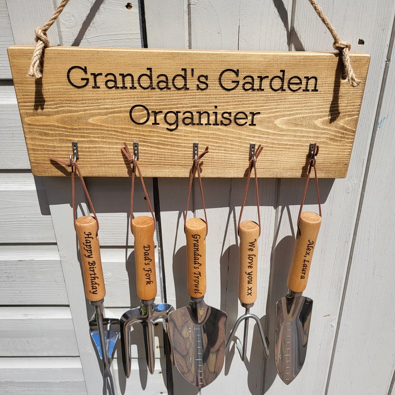 May include: Wooden garden organizer with the text "Grandad's Garden Organiser." It features five gardening tools with wooden handles and leather straps. The tools include a trowel, fork, and hand shovel, each with personalized engravings.