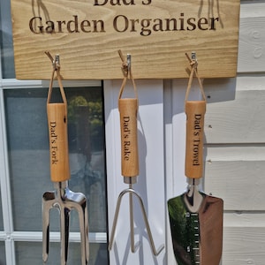 May include: A wooden sign that reads "Dad's Garden Organiser" with three garden tools hanging below. The tools include a fork, rake, and trowel, each with a wooden handle and the words "Dad's" and the tool name engraved on them.