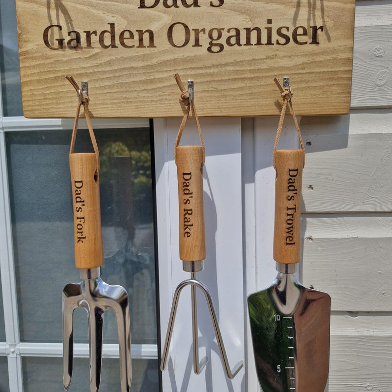 May include: A wooden sign that reads "Dad's Garden Organiser" with three garden tools hanging below. The tools include a fork, rake, and trowel, each with a wooden handle and the words "Dad's" and the tool name engraved on them.