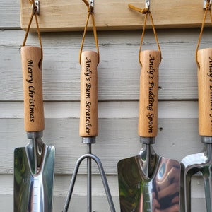 May include: A set of personalized garden tools with wooden handles and stainless steel blades. The handles are engraved with phrases like "Merry Christmas" and "Andy's Bum Scratcher." Each tool has a leather loop for hanging.