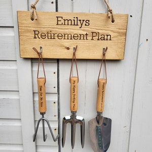 May include: A wooden sign with the text "Emilys Retirement Plan" is suspended by a rope. Below, three gardening tools hang from leather straps. The tools include a hand rake, a garden fork, and a trowel, each with wooden handles.