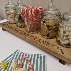 May include: A wooden serving tray with glass jars filled with candy. The tray is engraved with "CANDY CANE LANE". The jars contain various candies, including candy canes, gold-wrapped chocolate coins, and other sweets. A candy cane and sweets are on a striped napkin.
