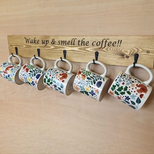 May include: A wooden coffee mug rack with five white ceramic mugs. Each mug has a floral design with red, blue, and green accents. The wooden rack has the text "Wake up & smell the coffee!!" in black.