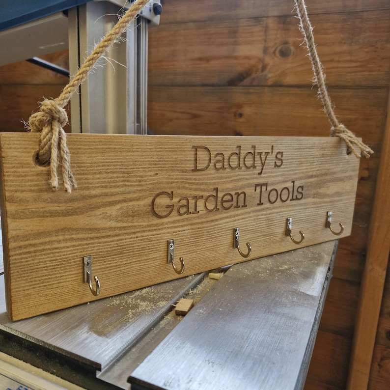 May include: Wooden sign with the text "Daddy's Garden Tools" engraved on it. The sign has six metal hooks and is suspended by a rope. The wood has a natural grain and a light brown color.