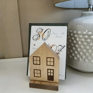 May include: A wooden house-shaped card holder with four windows and a door. Behind it is a card with the text "Save The Date" and floral accents. A decorative lamp is in the background.