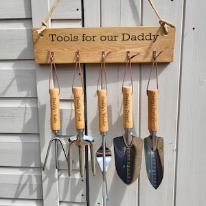 May include: A wooden sign with the text "Tools for our Daddy" hangs above a set of five gardening tools. Each tool has a wooden handle and a leather loop for hanging. The tools include a rake, fork, weeder, trowel, and transplanter.