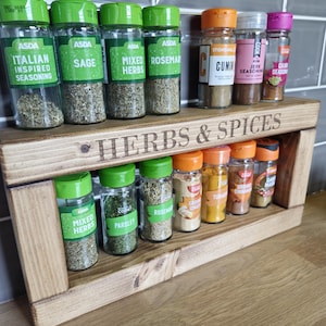 May include: A wooden spice rack labeled "HERBS & SPICES" holds various spice jars with green and orange lids. The jars contain herbs and spices like rosemary, cumin, and turmeric. The rack is made of light-colored wood and is displayed on a countertop.
